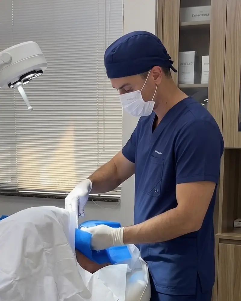 Dr. Luciano Ferreira usando uniforme cirúrgico azul, máscara e luvas brancas, focado enquanto realiza um procedimento clínico minucioso no rosto de um paciente em seu consultório médico.